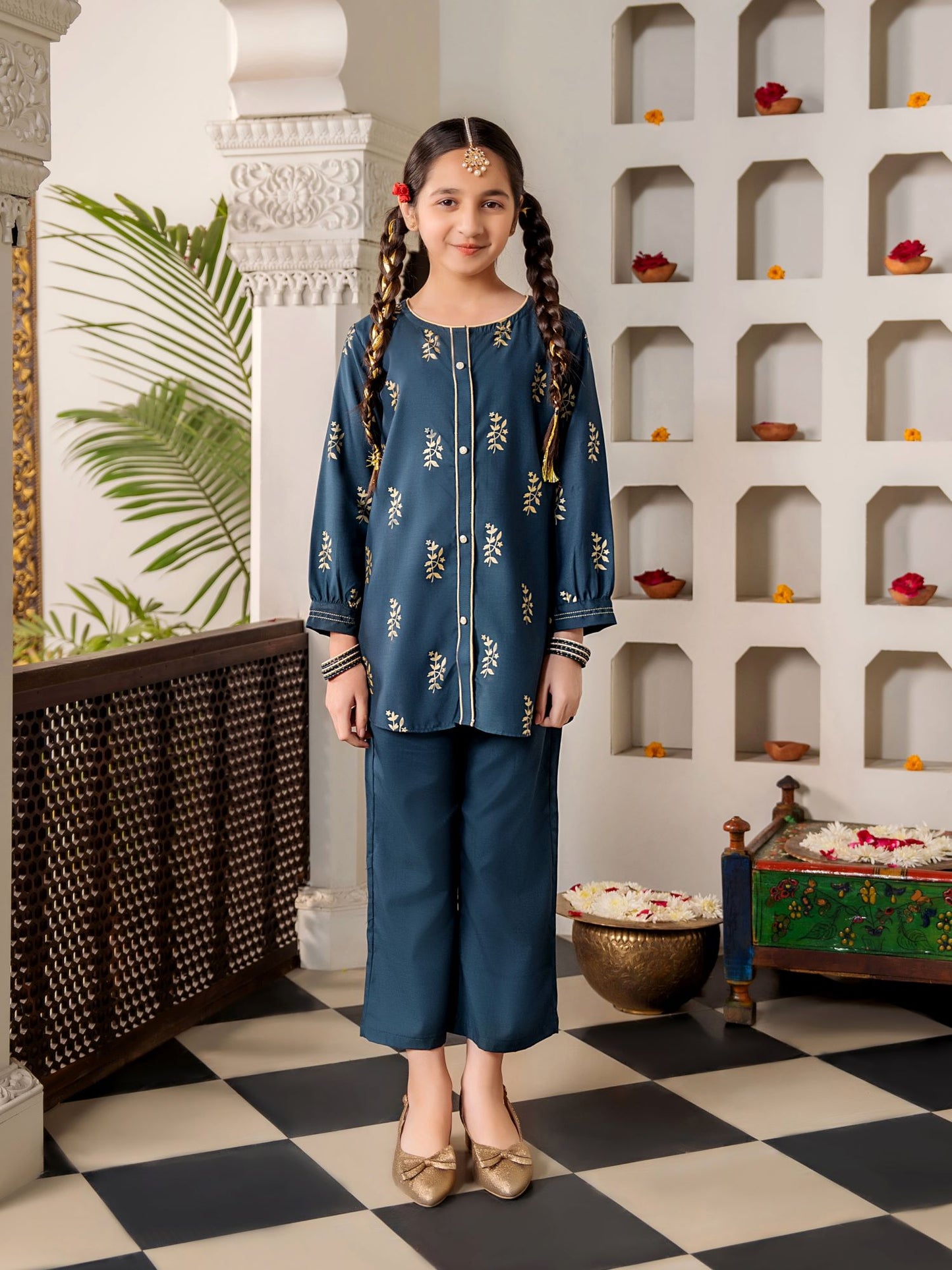 Young girl in a traditional blue outfit standing in a decorative room with shelves and plants.