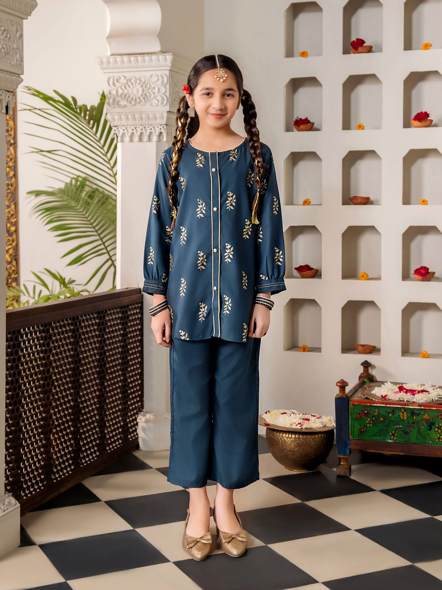 Young girl in a traditional blue outfit standing in a decorative room with shelves and plants.